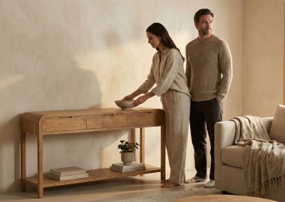 Koppel in woonkamer bij teak wandtafel Arus; vrouw legt schaal neer op de tafel met lade en plank, man staat ernaast, beige bank en vloerkleed.