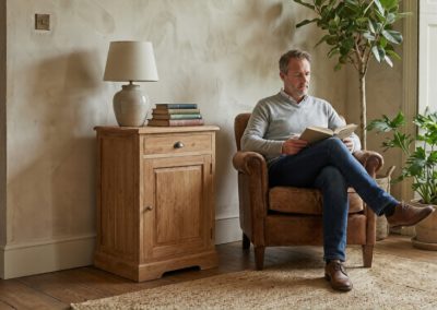 Man zit in fauteuil en leest naast teak dressoir Senja 1 deur met lamp en boeken, met plant op achtergrond.